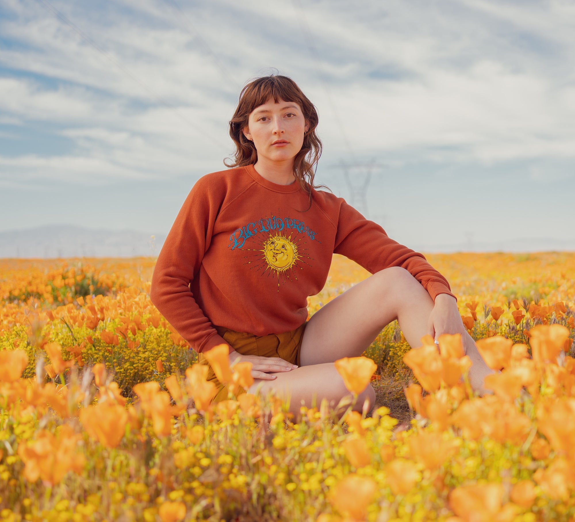 Alexandra Skye is sitting in a field of poppies wearing Bill Ogden's Sun Baby Crew and Work Shorts in Spicy Mustard
