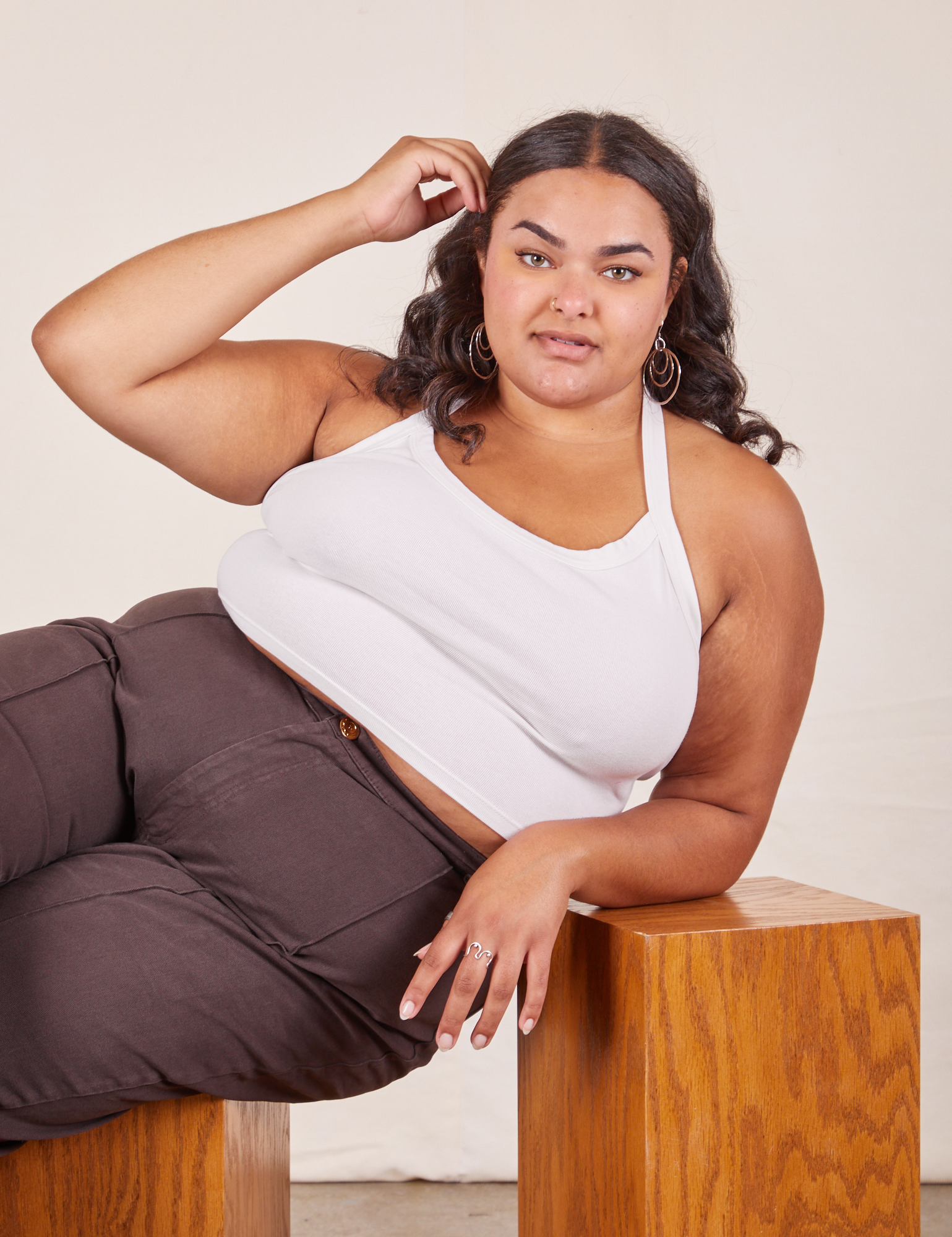 Alicia is 5'9" and wearing XL Halter Top in Vintage Tee Off-White paired with espresso brown Western Pants. She is sitting sideways on a wooden box and her right elbow is leaning on another wooden box.