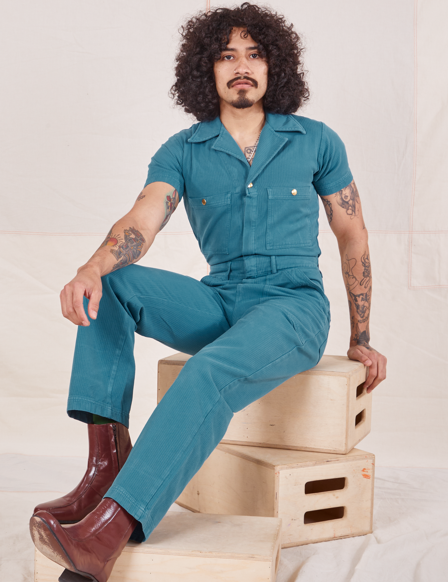 Jesse is 5&#39;8&quot; and wearing size S in Heritage Short Sleeve Jumpsuit in Marine Blue sitting on a stack of wooden crates