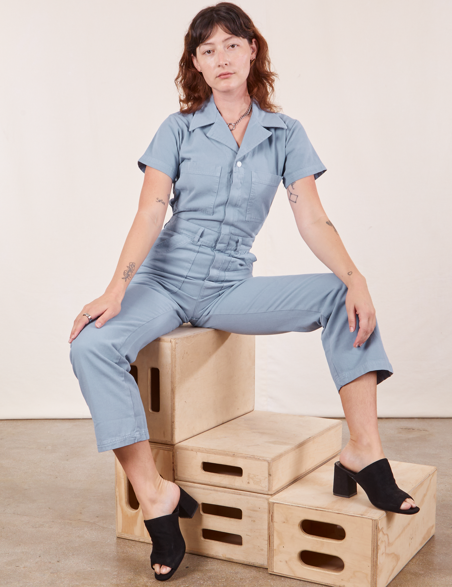Alex is wearing Short Sleeve Jumpsuit in Periwinkle and sitting on a stack of wooden crates