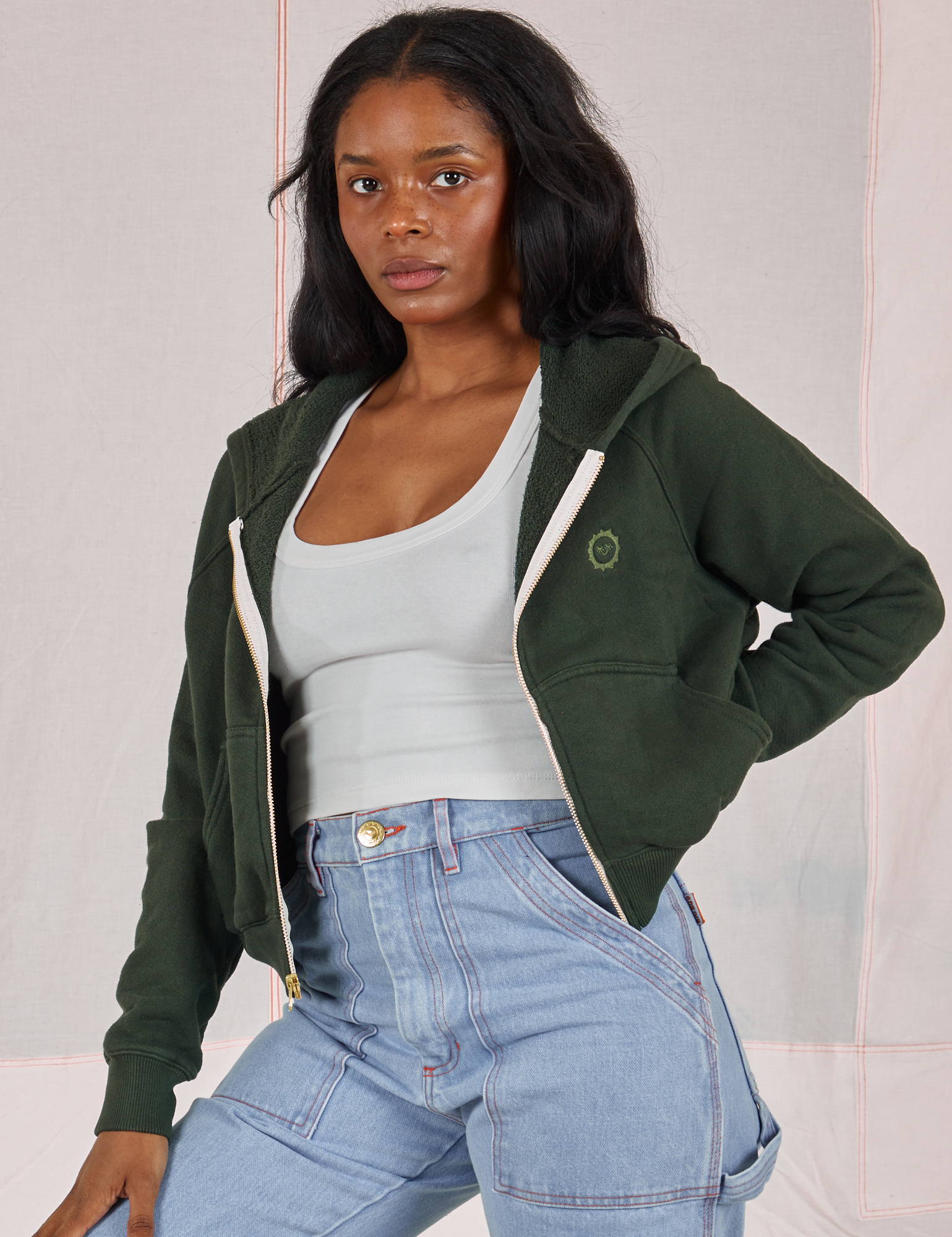 Kandia is 5'3" and wearing P Cropped Zip Hoodie in Swamp Green paired with a vintage off-white Cropped Tank underneath and light wash Carpenter Jeans