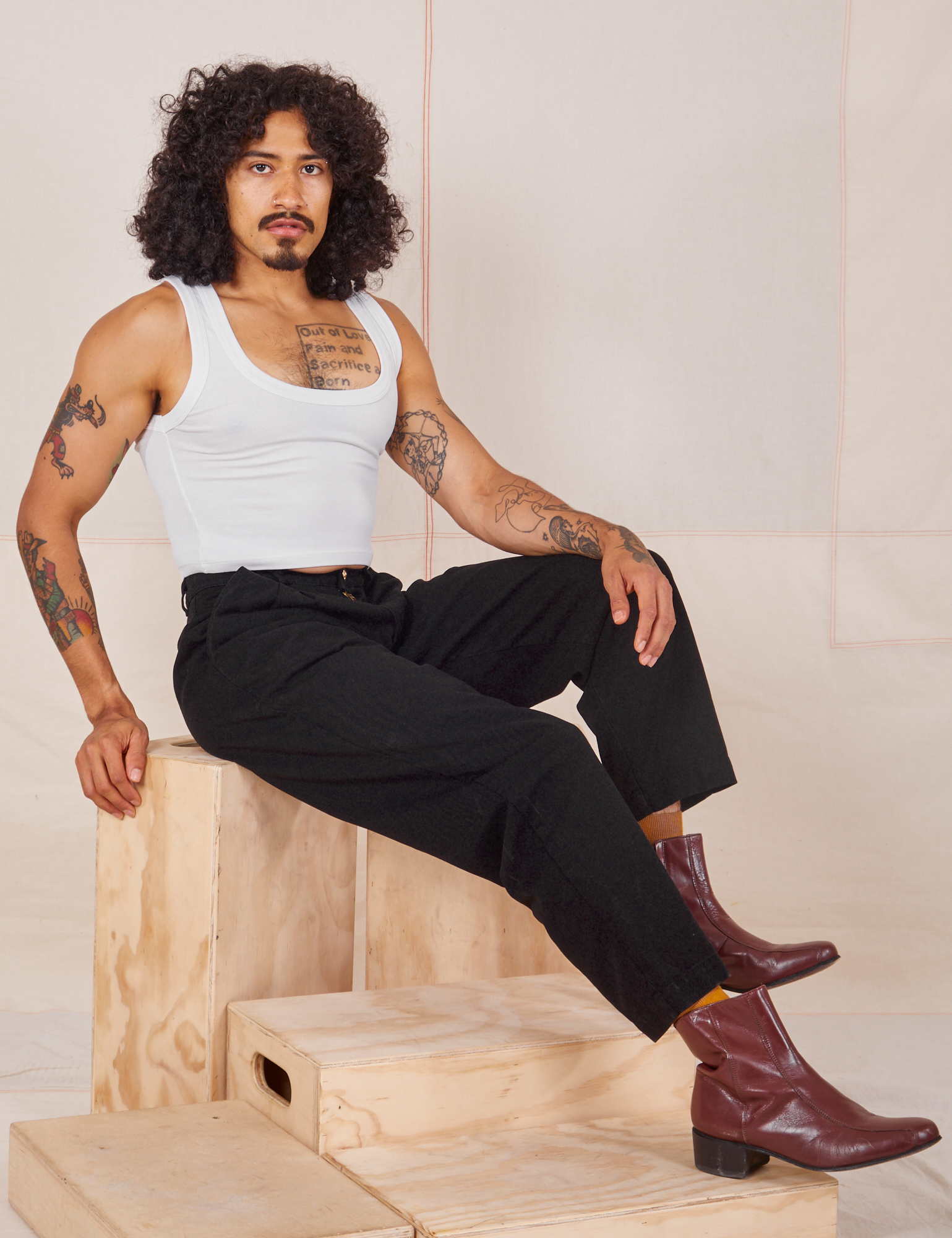Jesse is sitting on a wooden crate wearing Heavyweight Trousers in Basic Black and Cropped Tank Top in vintage tee off-white