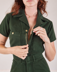 Heritage Short Sleeve Jumpsuit in Swamp Green front close up. Alex is pulling down the zipper.