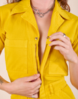 Short Sleeve Jumpsuit in Golden Yellow front close up. Alex is pulling down on the zipper.