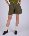 Close up of Pleated Trouser Shorts in Surplus Green with Alex's hands in both front pockets.