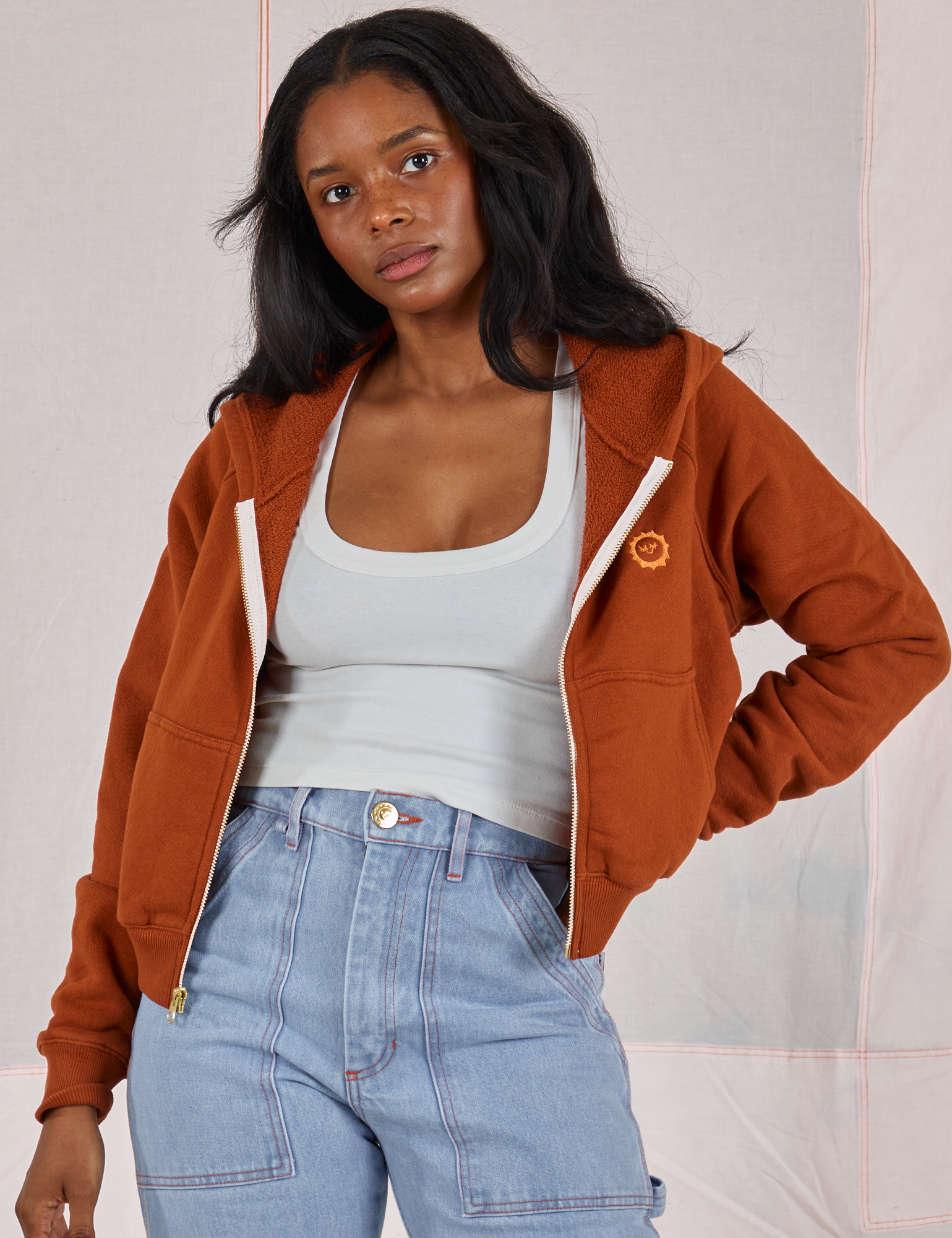 Kandia is wearing Cropped Zip Hoodie in Burnt Terracotta, vintage off-white Cropped Tank and light wash Carpenter Jeans