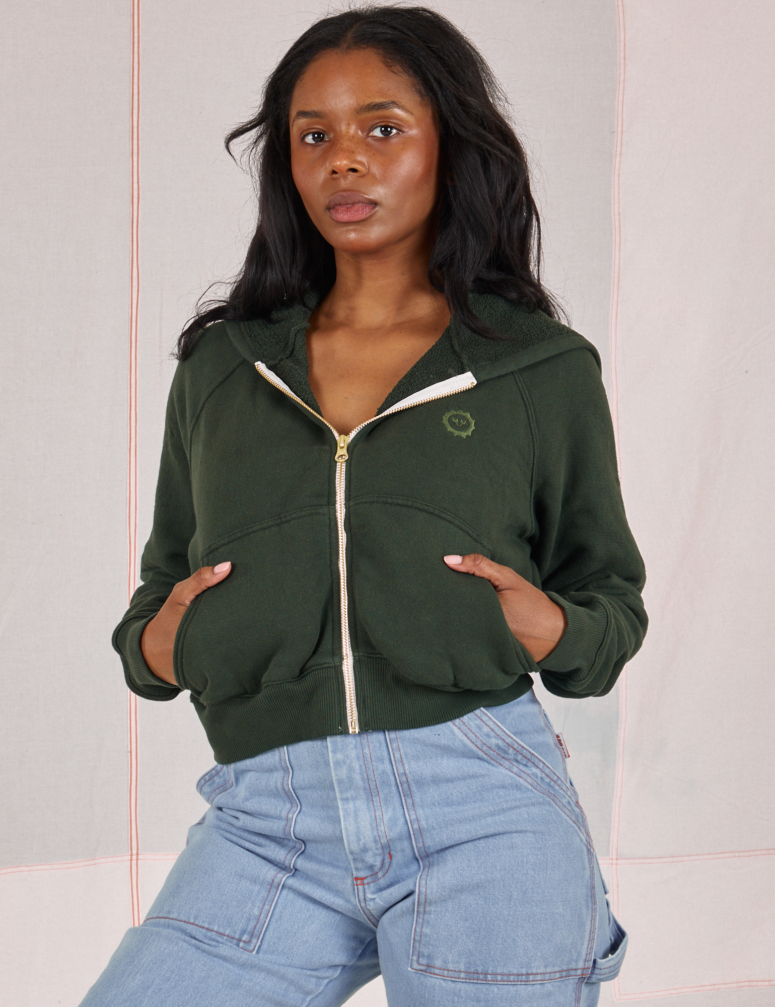 Kandia is wearing a zipped up Cropped Zip Hoodie in Swamp Green and light wash Carpenter Jeans