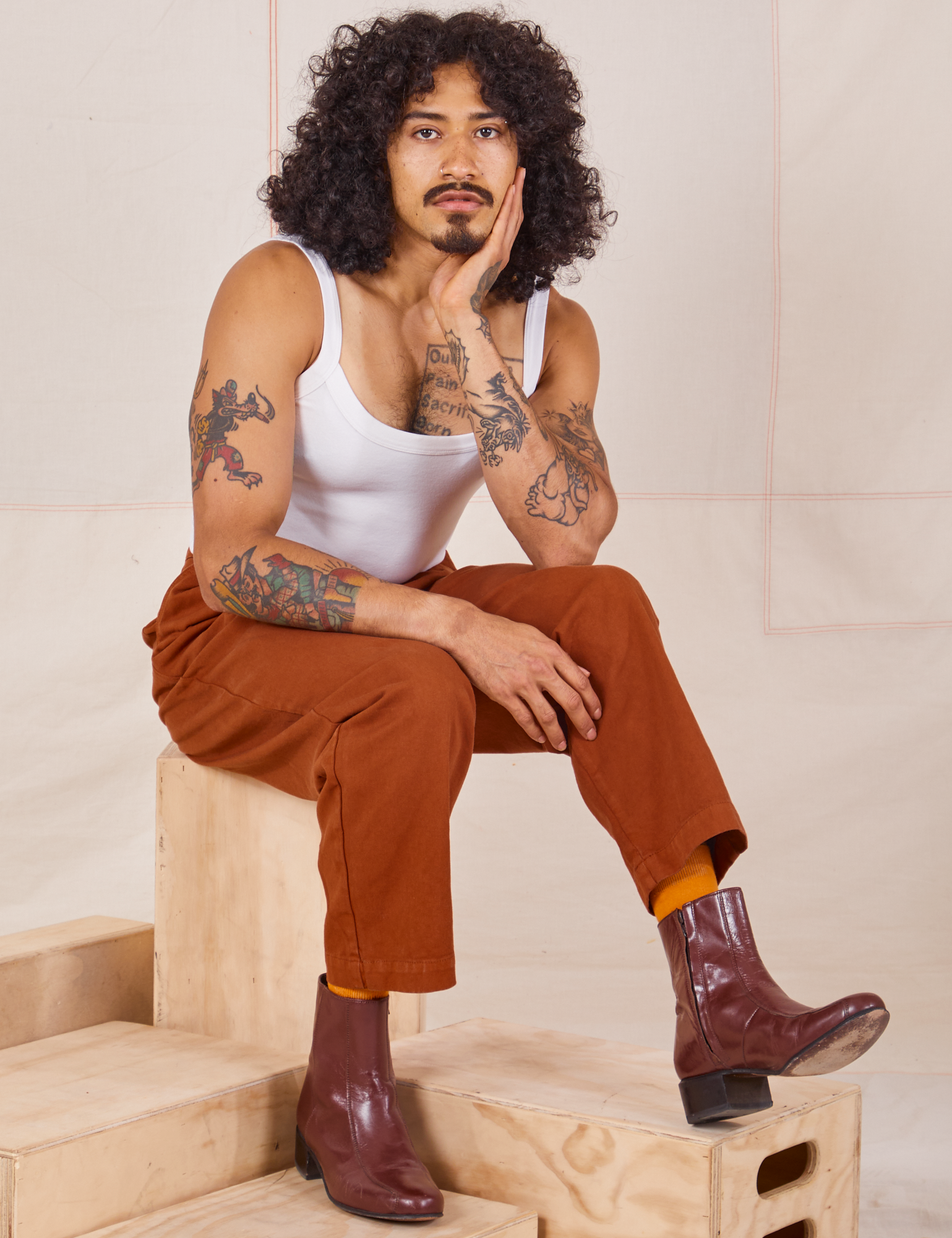 Jesse is sitting on a wooden crate. They are wearing Heavyweight Trousers in Burnt Terracotta and Cropped Cami in vintage tee off-white