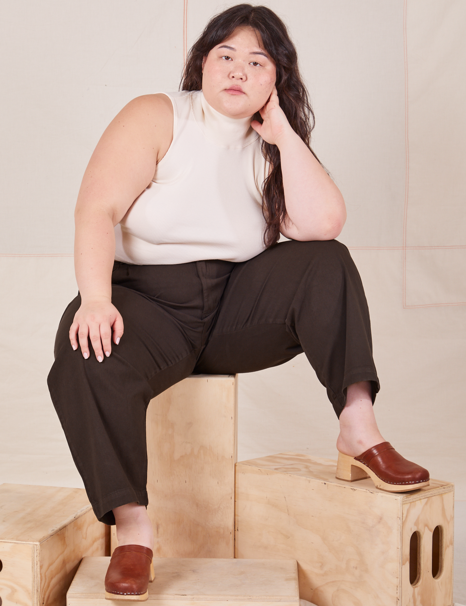 Ashley is sitting on a wooden crate wearing Heavyweight Trousers in Espresso Brown and Sleeveless Turtleneck in vintage tee off-white
