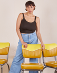 Tiara is standing behind a yellow chair with her hands resting on the back. She is wearing Cropped Cami in Espresso Brown and light wash Sailor Jeans
