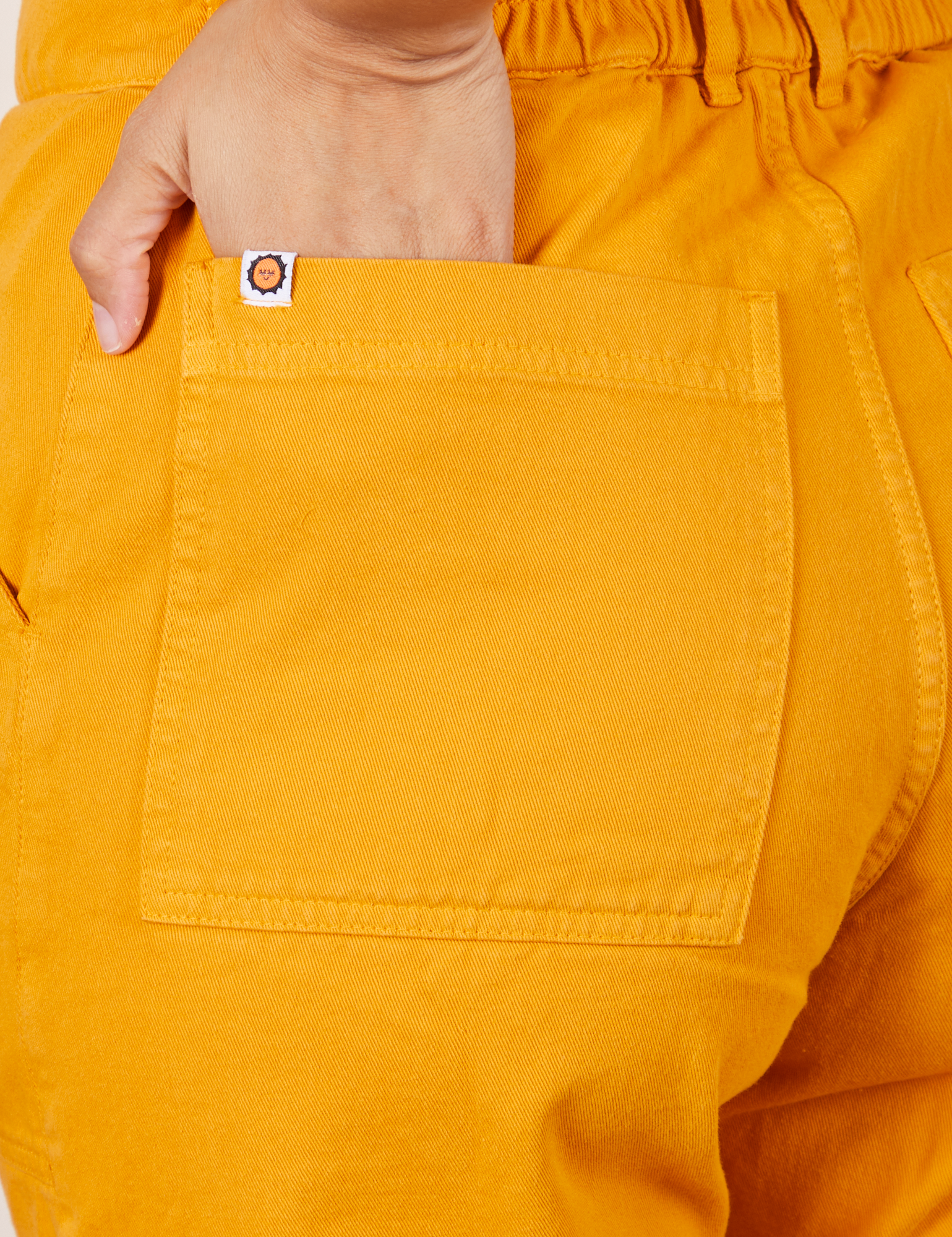 Back pocket close up of Short Sleeve Jumpsuit in Mustard Yellow. Tiara has her hand in the pocket.