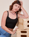 Alex is sitting on the ground and leaning on a stack of wooden crates. She is wearing Cropped Cami in Basic Black.