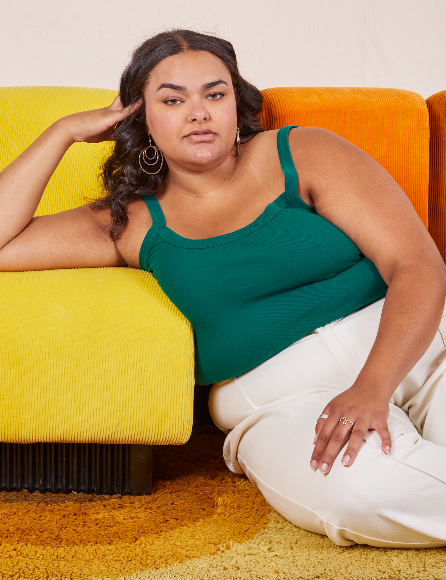 Alicia is sitting on a shaggy yellow rug with her right arm on a yellow upholstered chair. She is wearing Cropped Cami in Hunter Green and vintage tee off-white Western Pants.