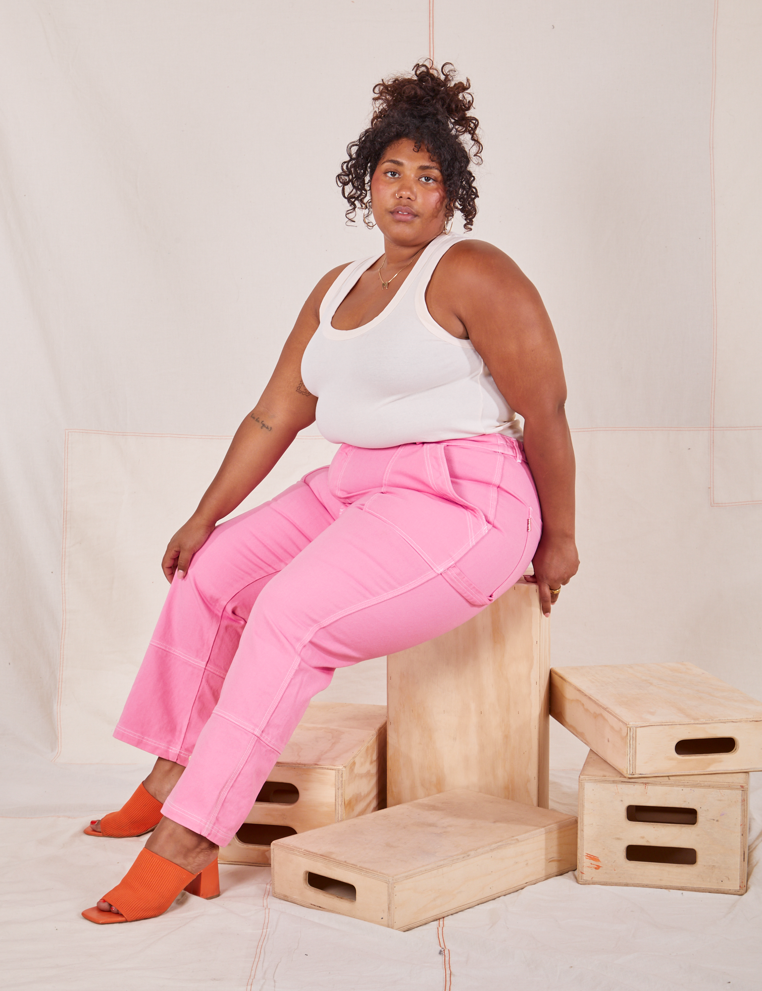 Morgan is sitting on a wooden crate wearing Carpenter Jeans in Bubblegum Pink and Tank Top in vintage tee off-white