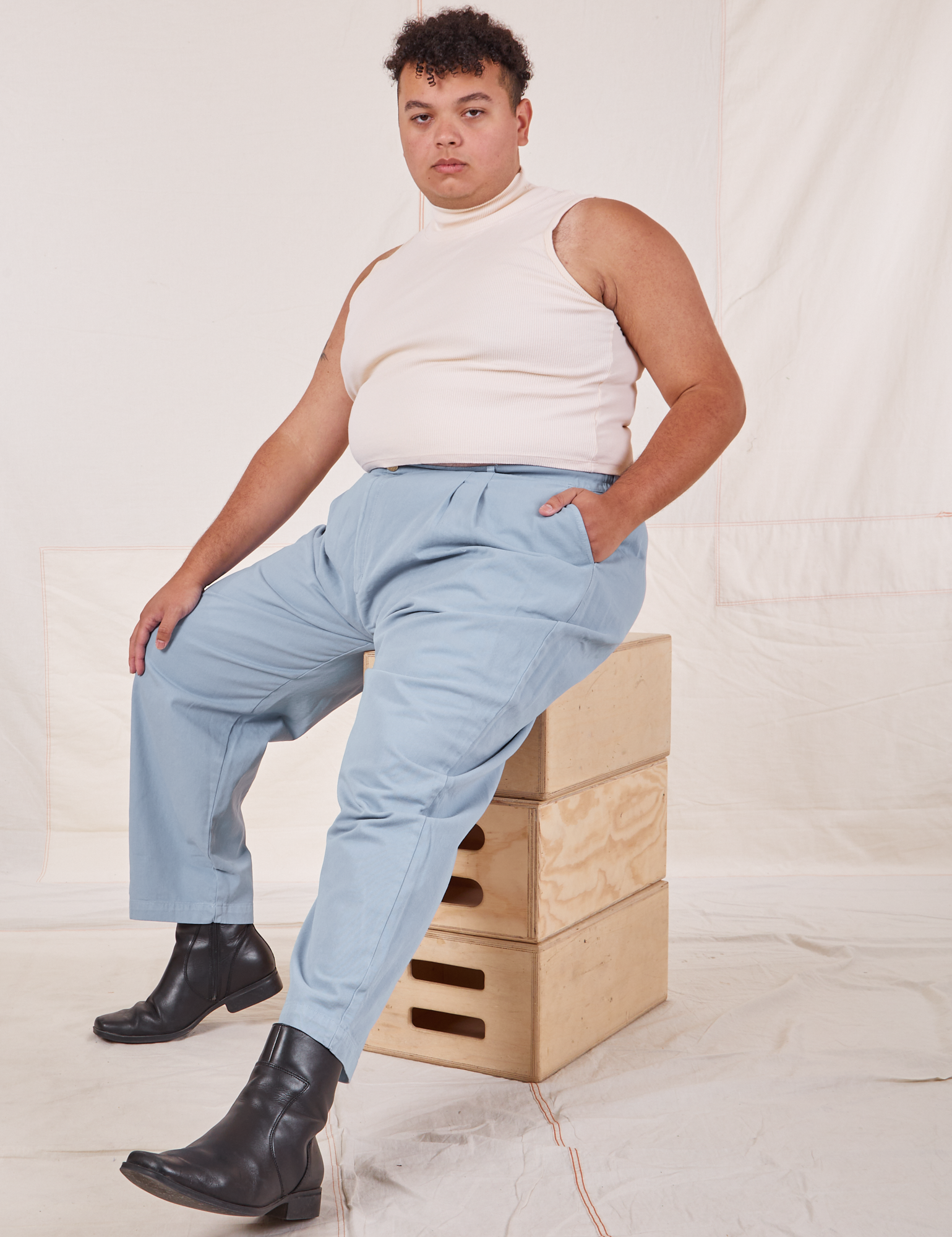 Miguel is wearing Heavyweight Trousers in Periwinkle and vintage off-white Sleeveless Tank Top. He is sitting on a stack of wooden crates.