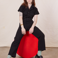 Hana is sitting on a red circular stand wearing Petite Short Sleeve Jumpsuit in Basic Black