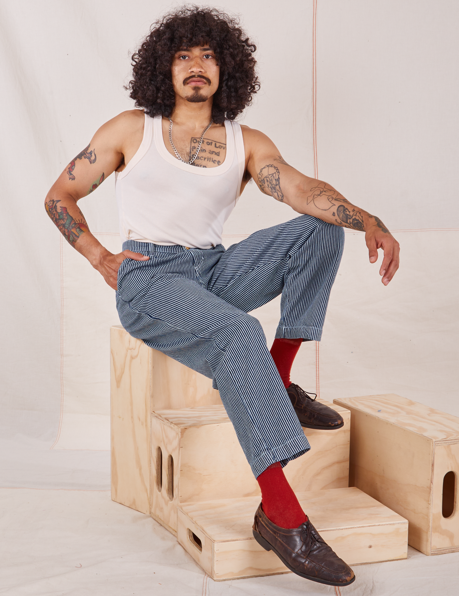 Jesse is sitting on a wooden crate wearing Denim Trouser Jeans in Railroad Stripe and Tank Top in vintage tee off-white
