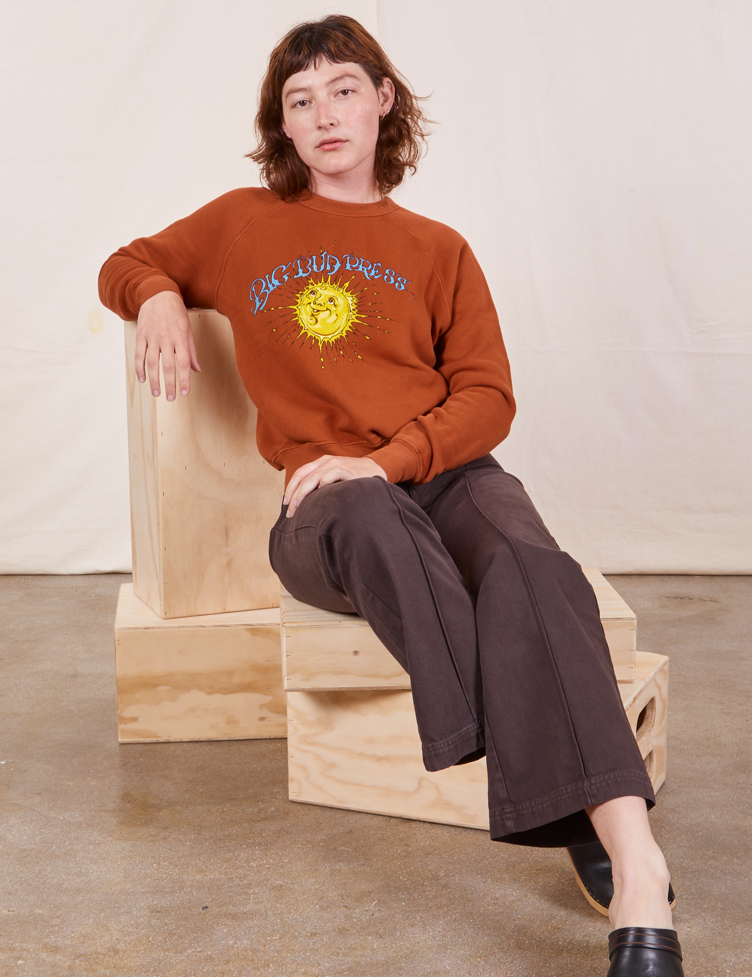 Alex is sitting on a wooden crate wearing Bill Ogden&#39;s Sun Baby Crew and espresso brown Western Pants