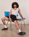 Jesse is sitting in a vintage brass and blue chair wearing Trouser Shorts in Basic Black and a vintage off-white Tank Top