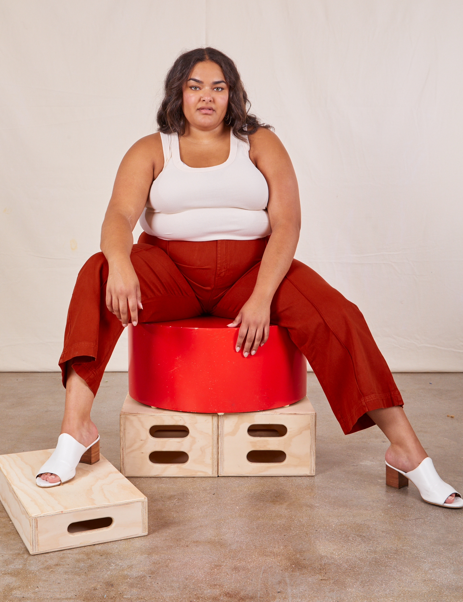 Alicia is sitting on a circular platform wearing Western Pants in Paprika and a Tank Top in vintage tee off-white