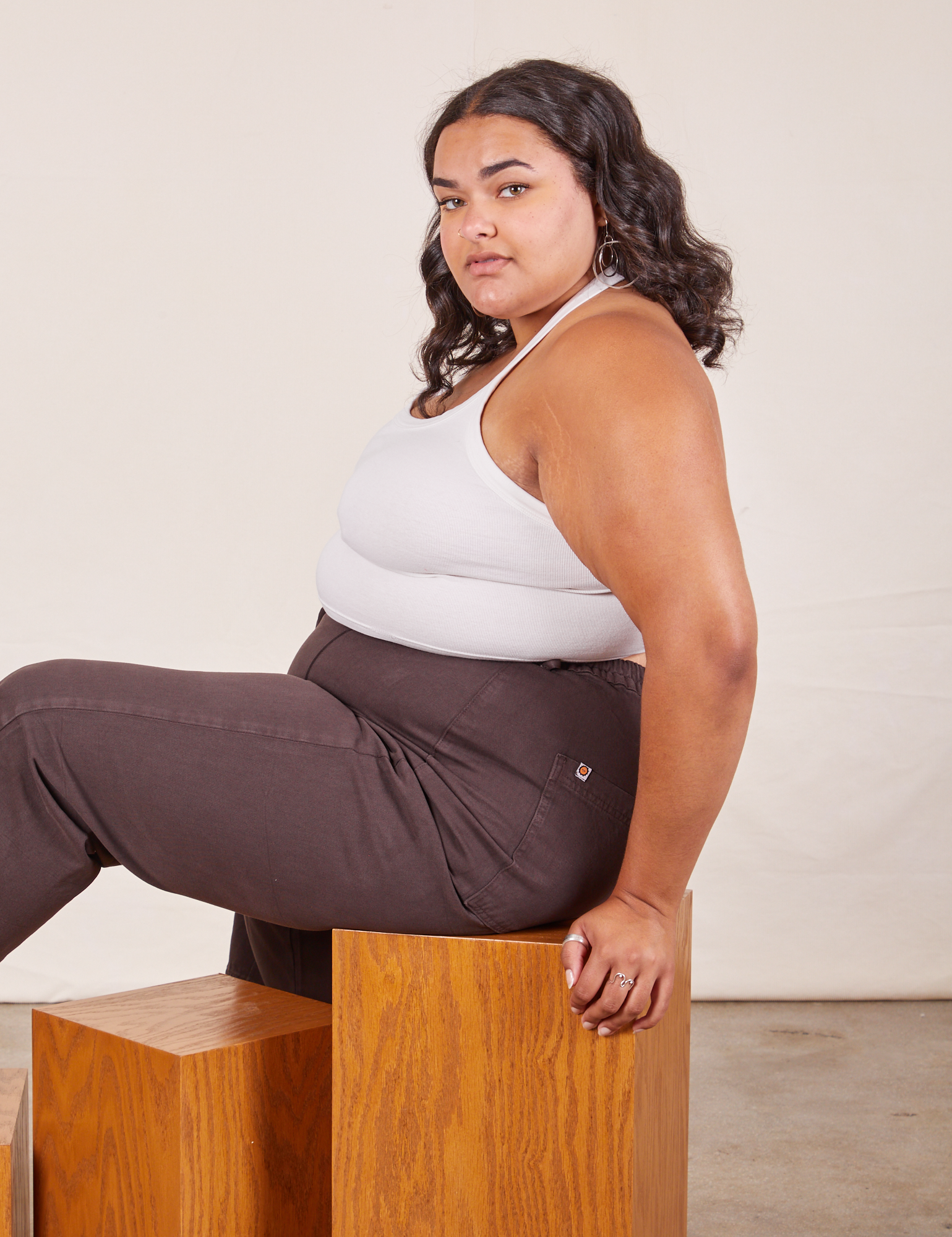 Alicia is sitting on a wooden box facing the left. She is wearing Halter Top in Vintage Tee Off-White and espresso brown Western Pants
