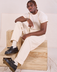 Issac is sitting on a wooden crate. He is wearing Heritage Trousers in Natural and Organic Vintage Tee in vintage tee off-white