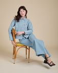 Alex is sitting on a chair and wearing Indigo Denim Work Jacket in Light Wash and light wash Indigo Wide Leg Trousers
