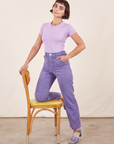 Work Pants in Faded Grape on Soraya wearing lilac purple Baby Tee. One knee is on a wooden chair with the other leg standing on the ground.