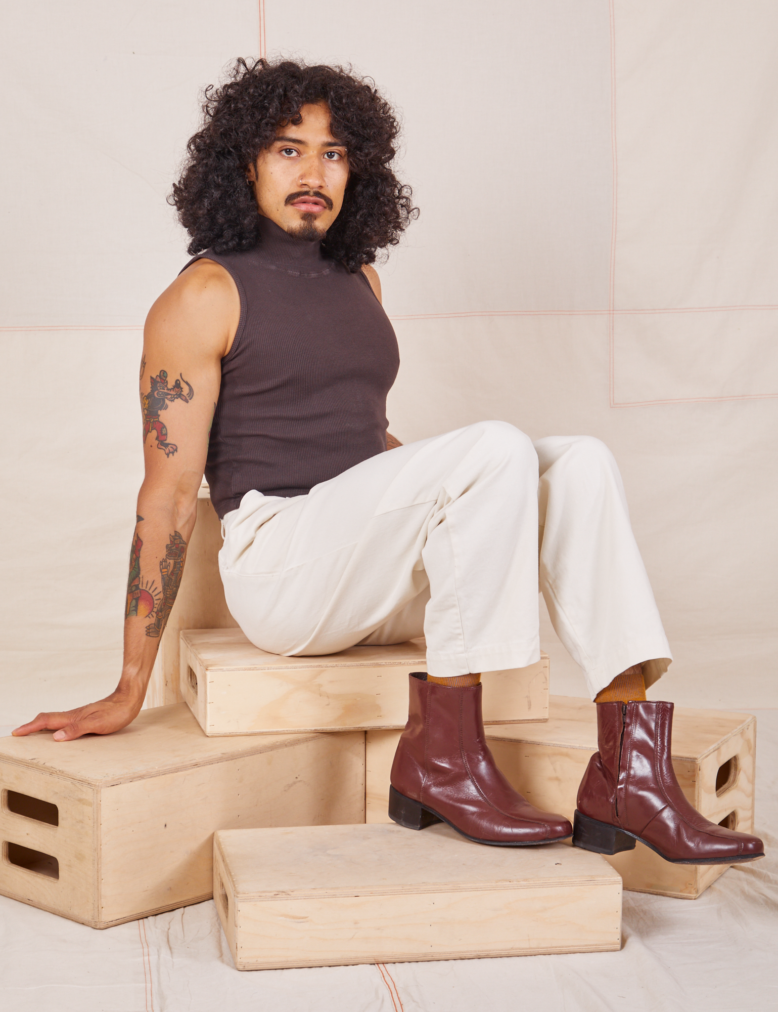 Jesse is sitting on a stack of wooden crates. They are wearing Heavyweight Trousers in Vintage Tee Off-White and espresso brown Sleeveless Turtleneck.