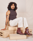 Jesse is sitting on a stack of wooden crates. They are wearing Heavyweight Trousers in Vintage Tee Off-White and espresso brown Sleeveless Turtleneck.