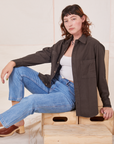 Alex is sitting on a wooden crate. She is wearing Oversize Overshirt in Espresso Brown, vintage off-white Tank Top and light wash Frontier Jeans