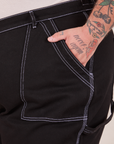 Front pocket close up of Carpenter Jeans in Black. Contrast white top stitching along edges of pocket. Sam has their hand in the pocket.