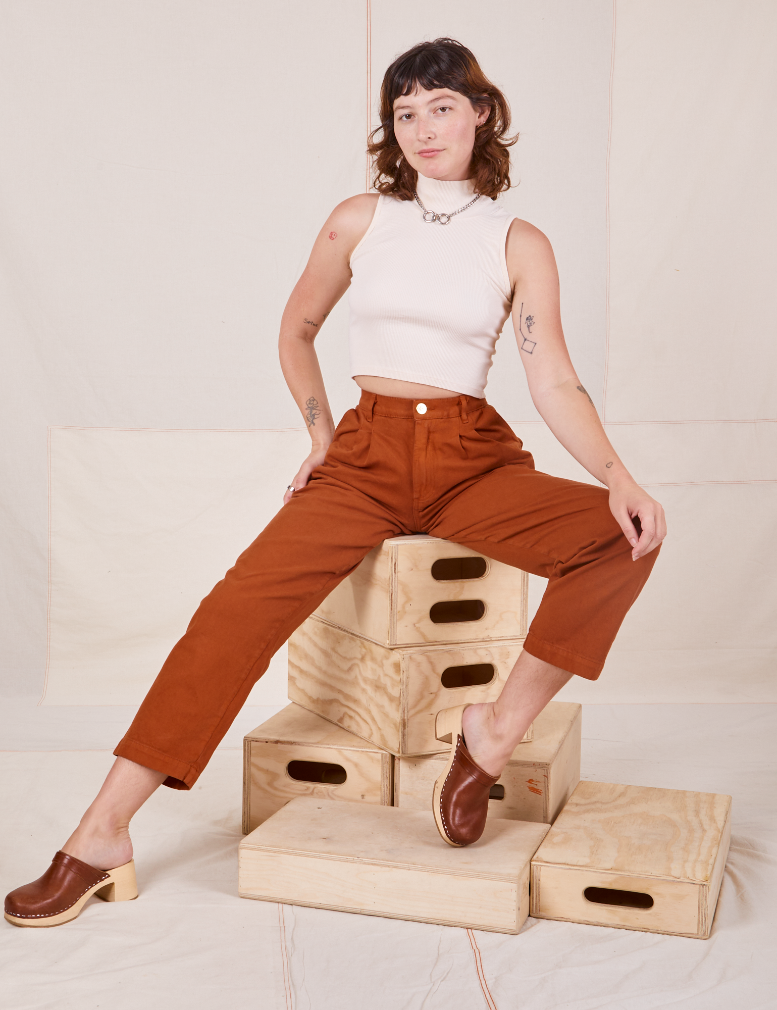 Alex is sitting on a stack of wooden crates. She is wearing Heavyweight Trousers in Burnt Terracotta and Sleeveless Turtleneck in vintage tee off-white
