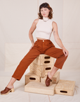 Alex is sitting on a stack of wooden crates. She is wearing Heavyweight Trousers in Burnt Terracotta and Sleeveless Turtleneck in vintage tee off-white