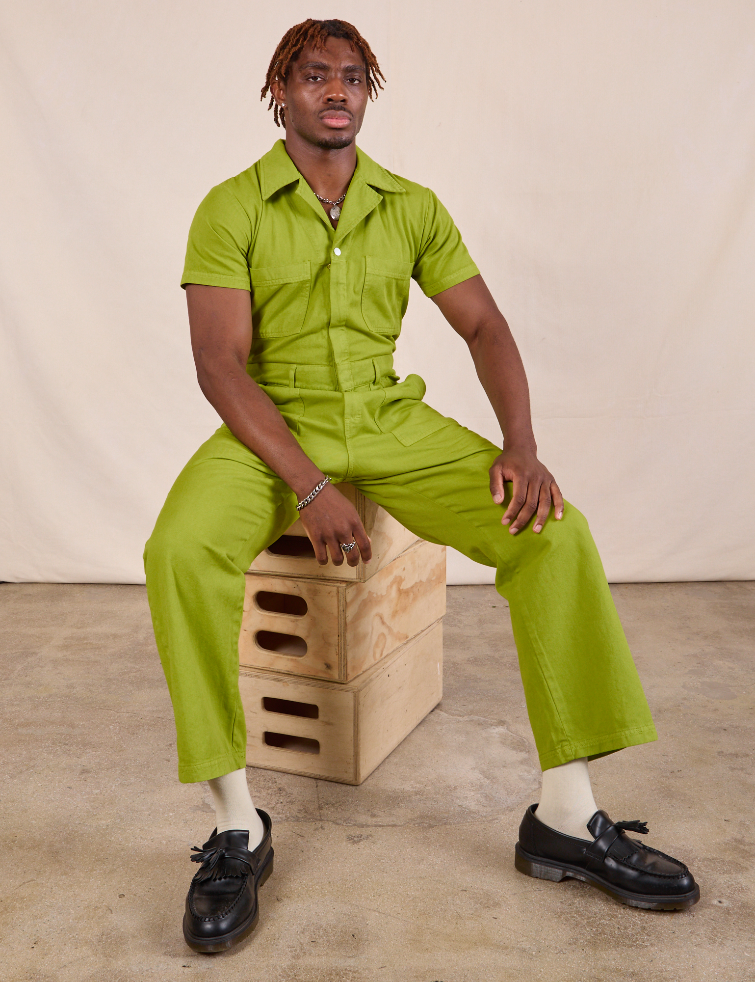 Issac is wearing Short Sleeve Jumpsuit in Gross Green and sitting on a stack of wooden crates