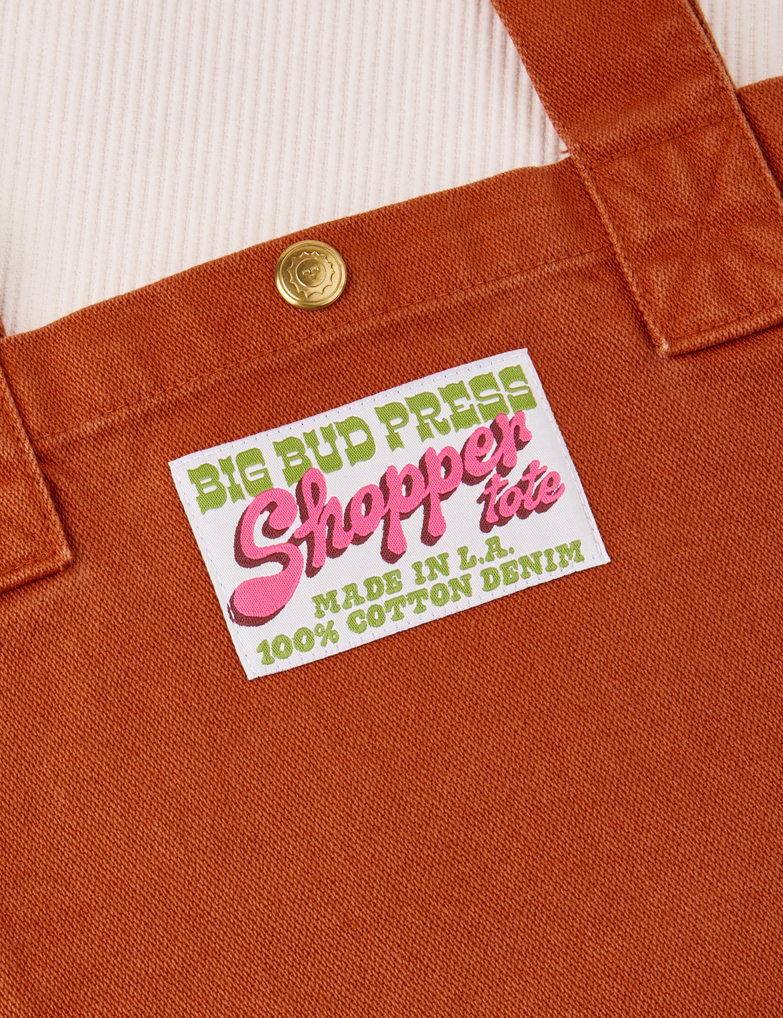 Close up of sun baby brass snap on Burnt Terracotta Shopper Tote Bag. Bag label in green and pink text that reads &quot;Big Bud Press Shopper Tote. Made in L.A, 100% Cotton Denim on a white background