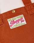Close up of sun baby brass snap on Burnt Terracotta Shopper Tote Bag. Bag label in green and pink text that reads "Big Bud Press Shopper Tote. Made in L.A, 100% Cotton Denim on a white background