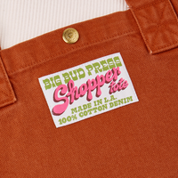 Close up of sun baby brass snap on Burnt Terracotta Shopper Tote Bag. Bag label in green and pink text that reads "Big Bud Press Shopper Tote. Made in L.A, 100% Cotton Denim on a white background
