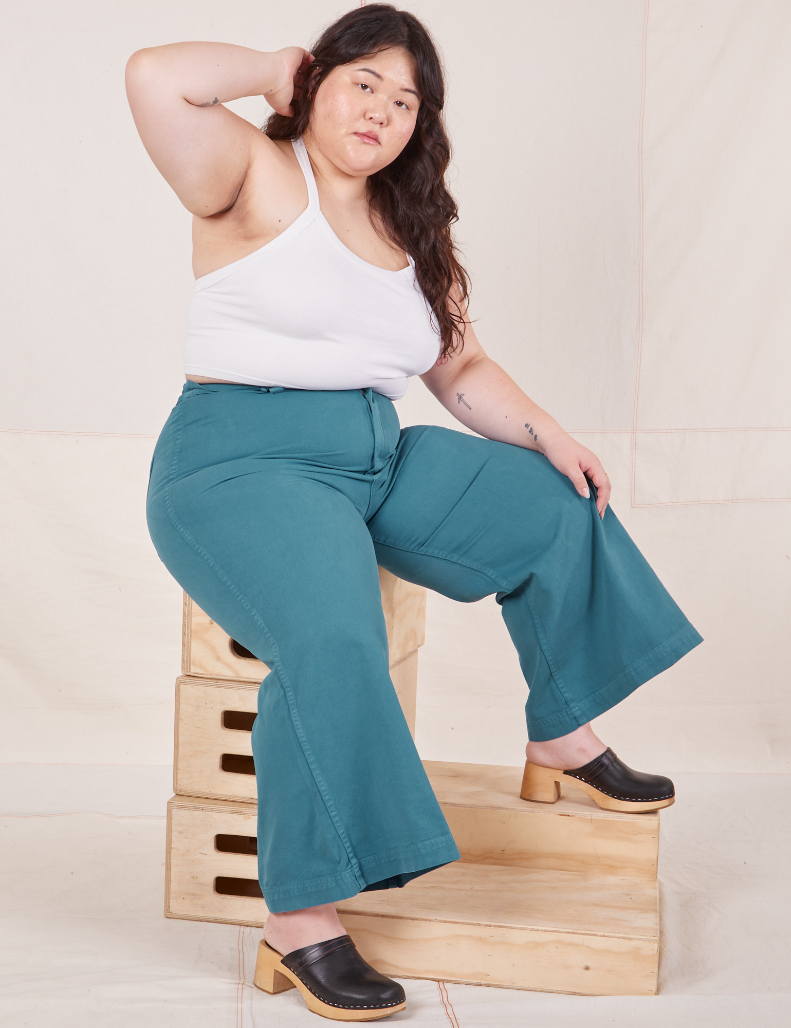 Ashley is sitting on a stack of wooden crates. She is wearing Petite Bell Bottoms in Marine Blue and Halter Top in vintage tee off-white