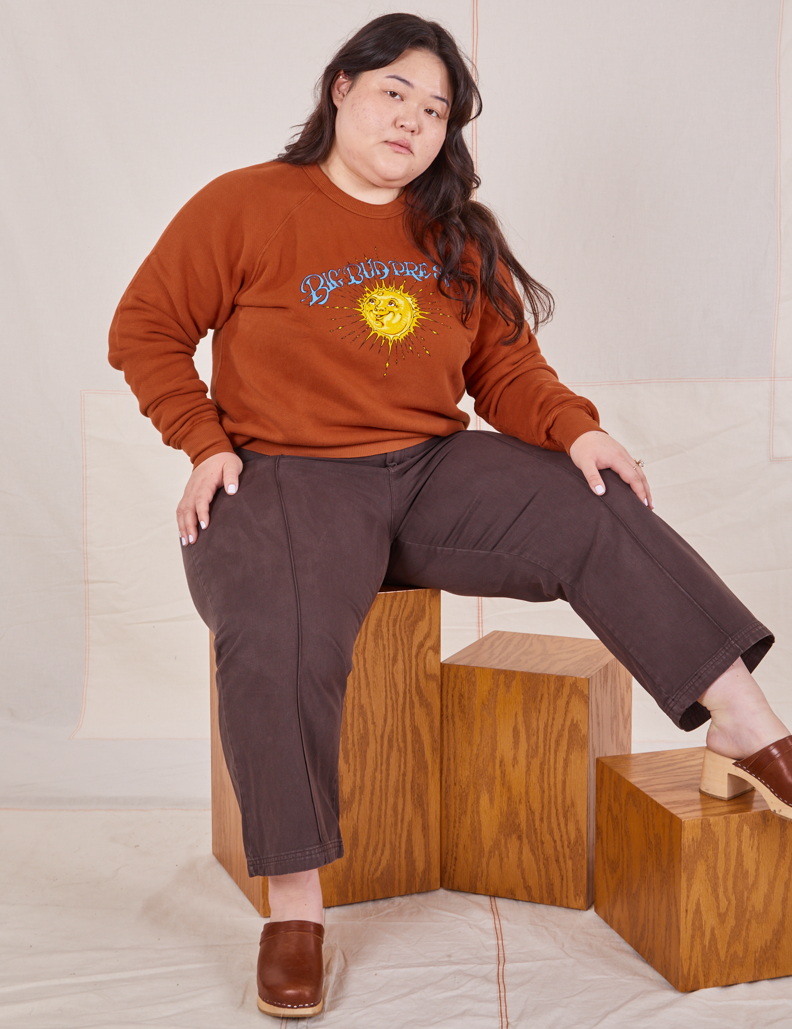 Ashley is sitting on a wooden crate wearing Bill Ogden&#39;s Sun Baby Crew and espresso brown Western Pants