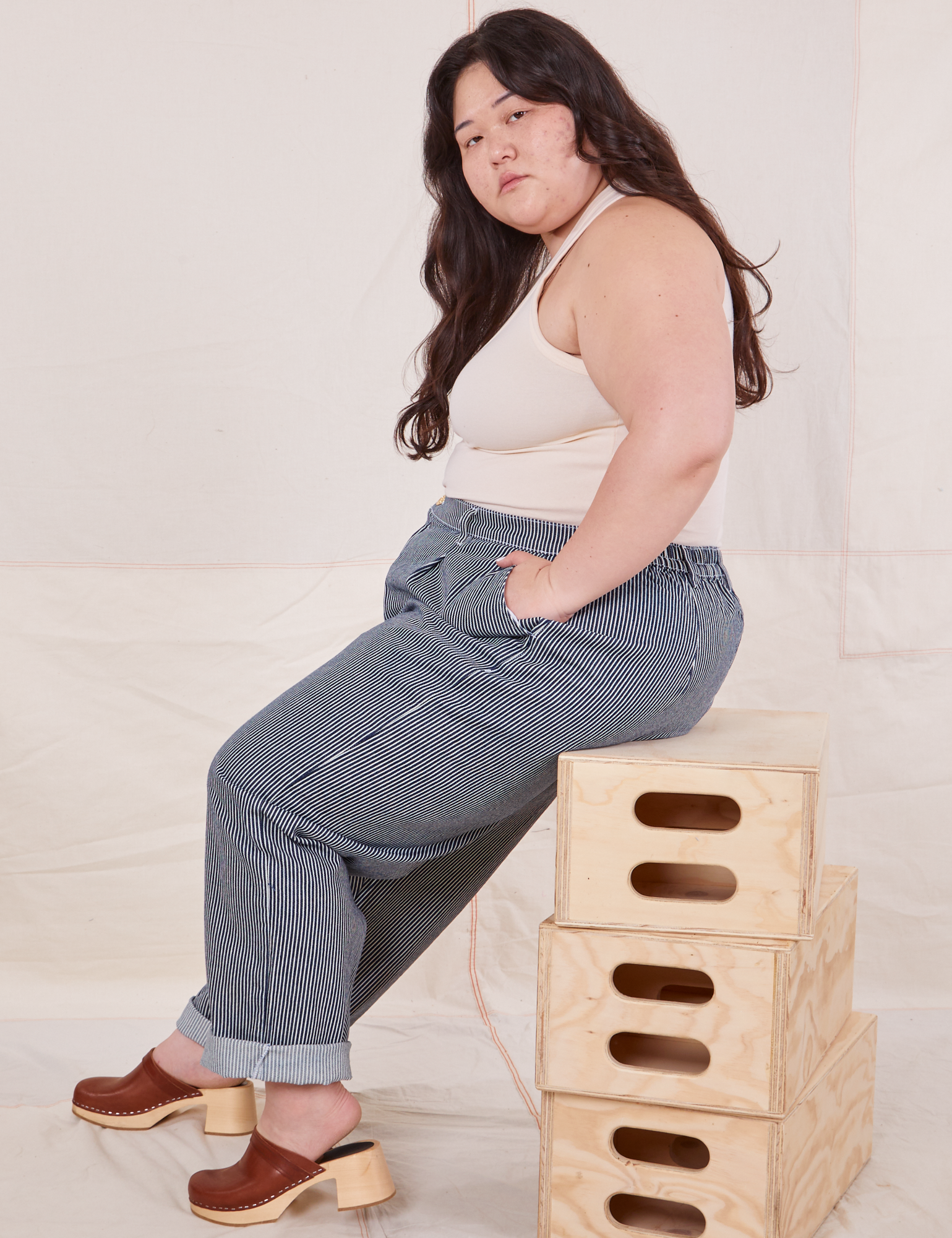 Ashley is sitting sideways on a stack of wooden crates. She is wearing Denim Trouser Jeans in Railroad Stripe and Tank Top in vintage tee off-white