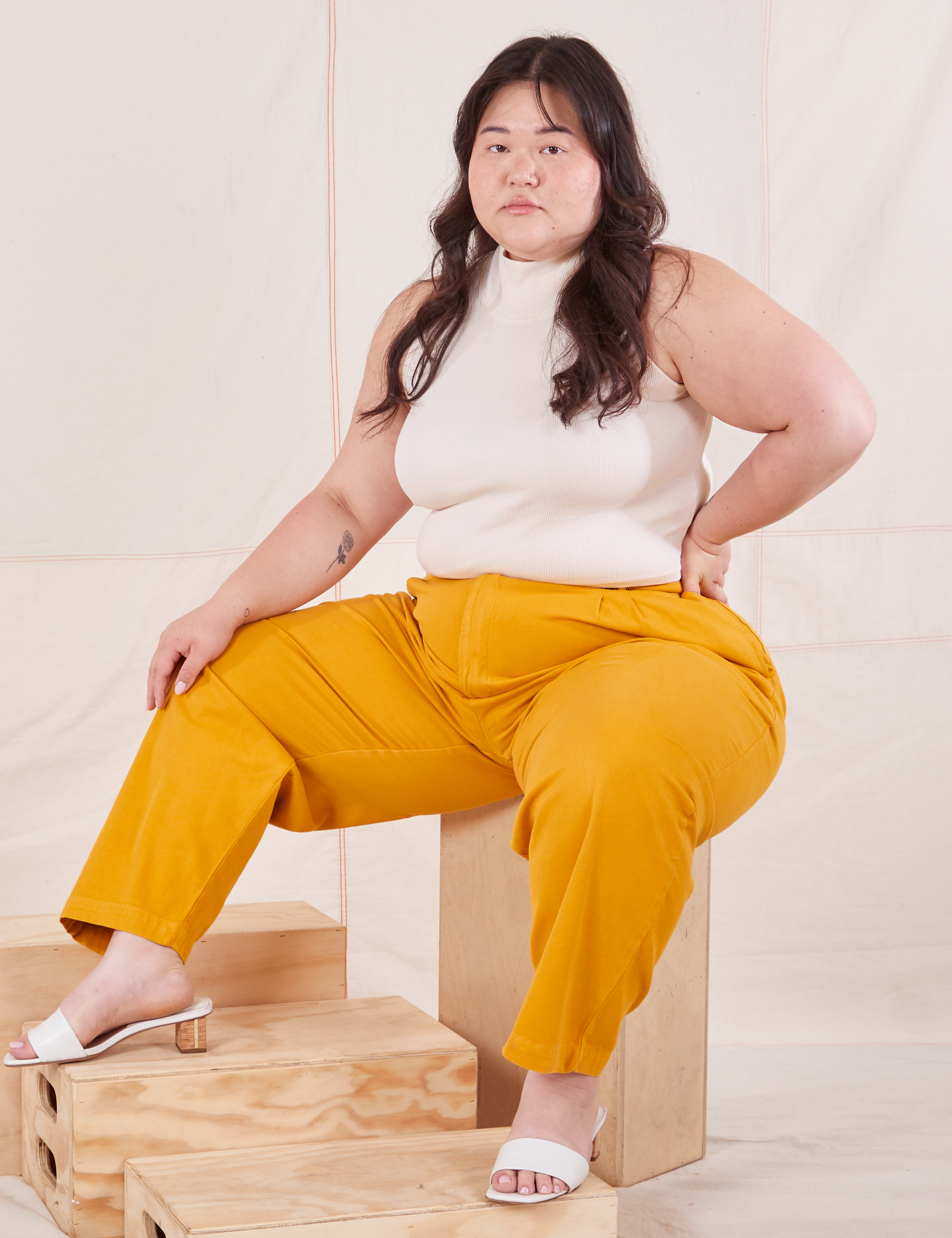 Ashley is sitting on a wooden crate wearing Organic Trousers in Mustard Yellow and Sleeveless Turtleneck in vintage tee off-white