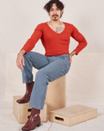 Jesse is sitting on a wooden crate wearing Railroad Stripe Denim Work Pants and a paprika Long Sleeve V-Neck Tee