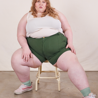 Catie is sitting on a vintage stool wearing Classic Work Shorts in Dark Emerald Green paired with a vintage off-white Tank Top