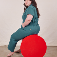 Ashley is sitting on a red circular platform wearing Petite Short Sleeve Jumpsuit in Marine Blue