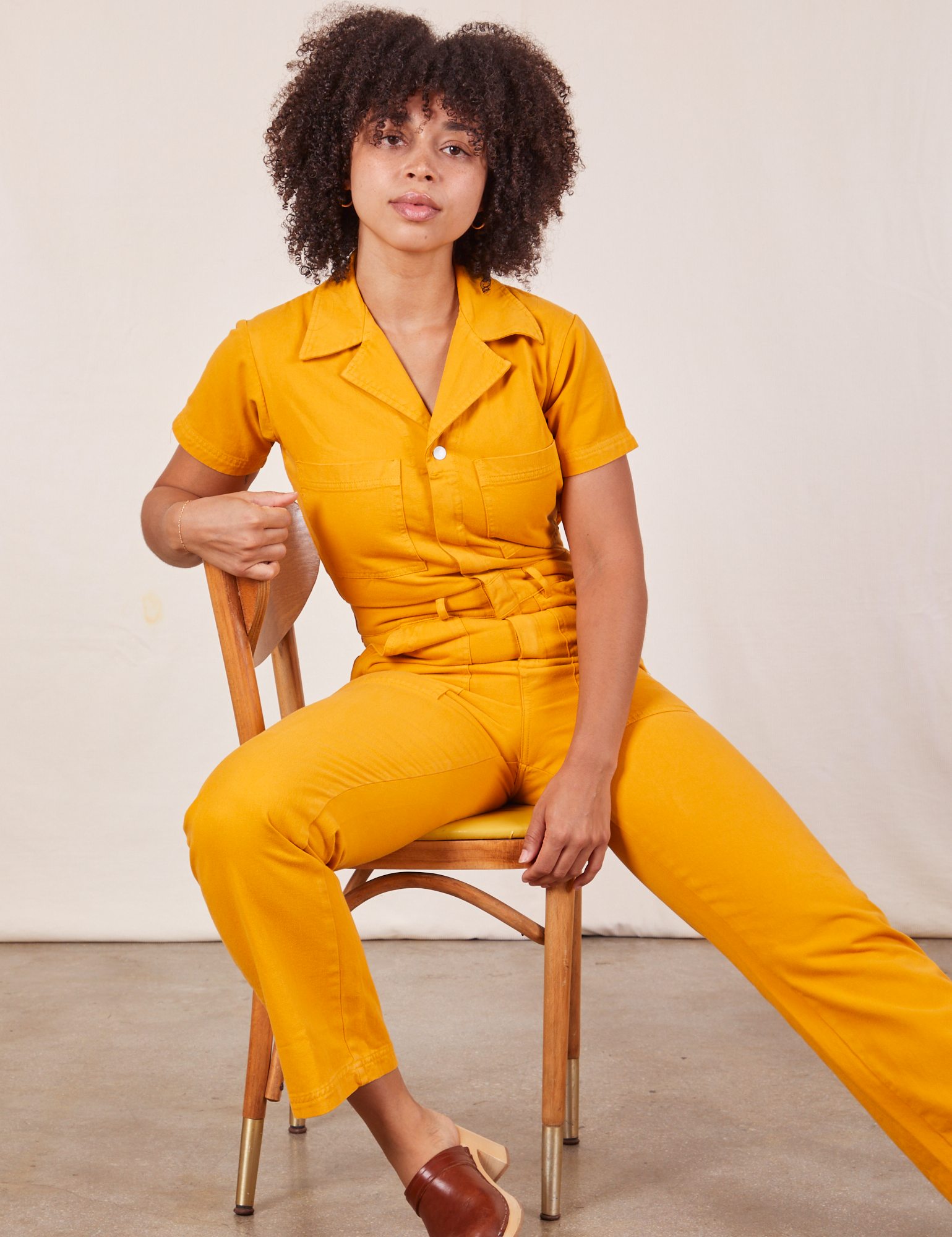 Gabi is sitting in a chair wearing Short Sleeve Jumpsuit in Mustard Yellow