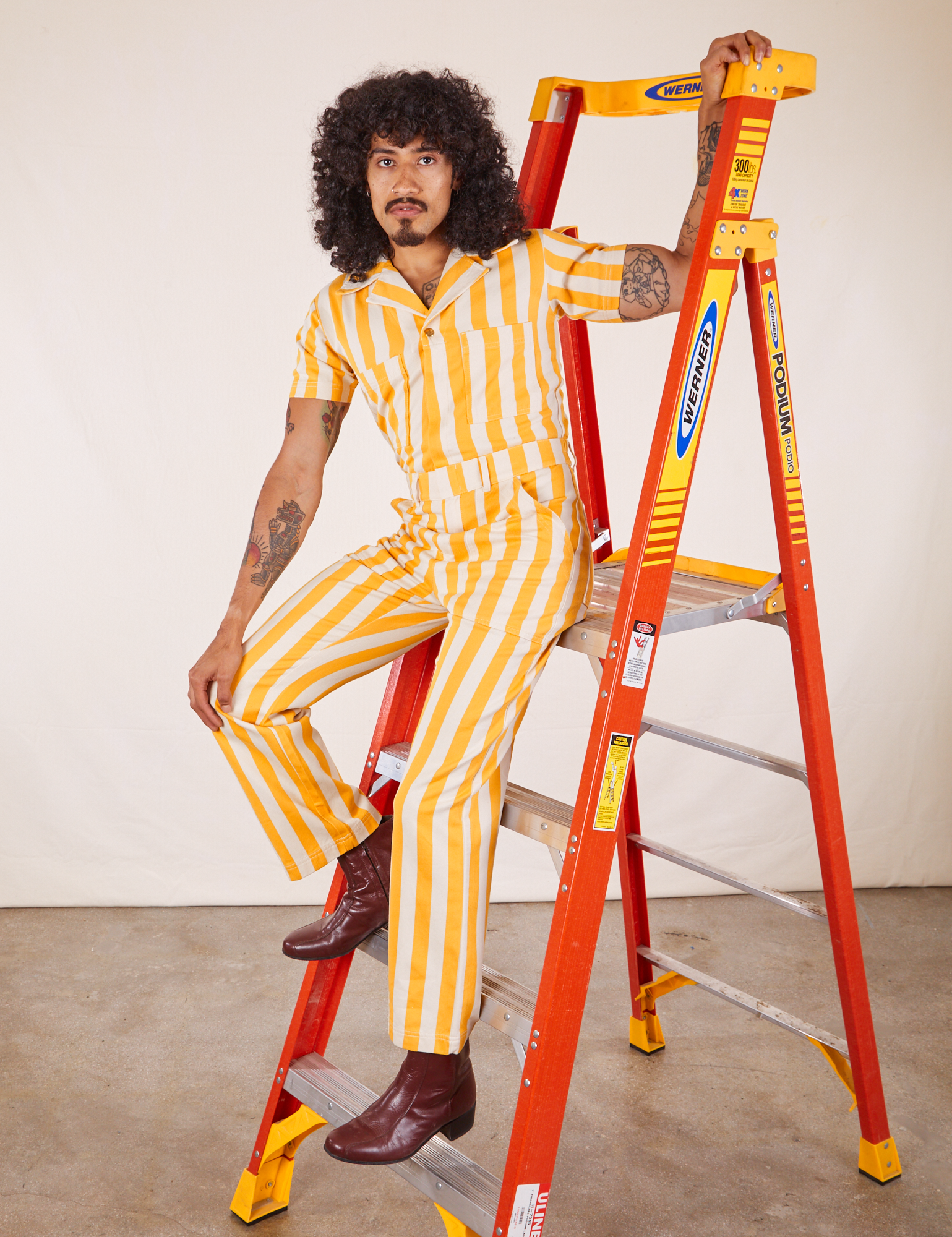 Jesse is sitting on a ladder. They are wearing Lemon Stripe Jumpsuit