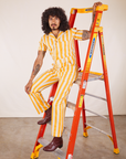 Jesse is sitting on a ladder. They are wearing Lemon Stripe Jumpsuit