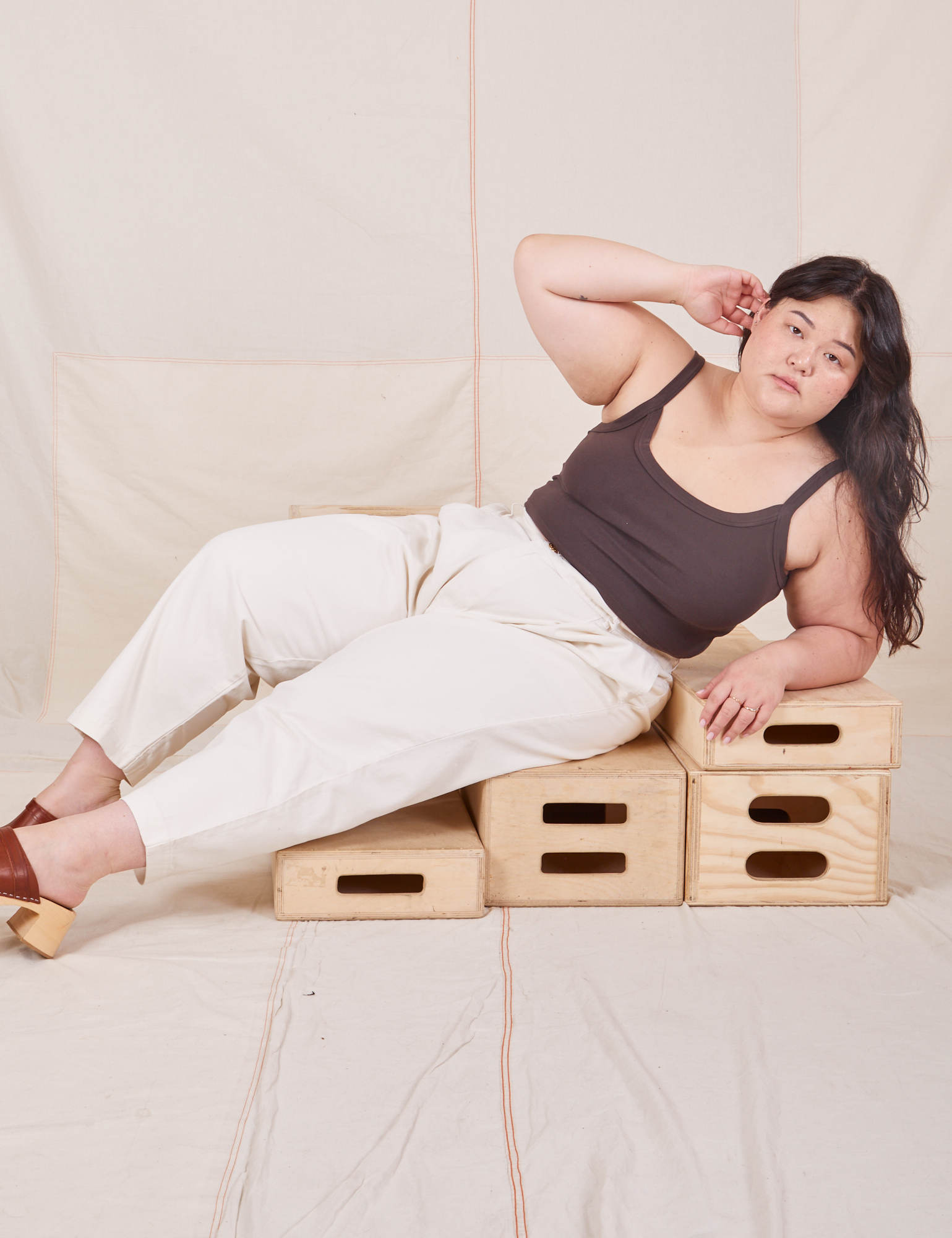 Ashley is lying across wooden crates wearing Heavyweight Trousers in Vintage Tee Off-White and espresso brown Cropped Cami.