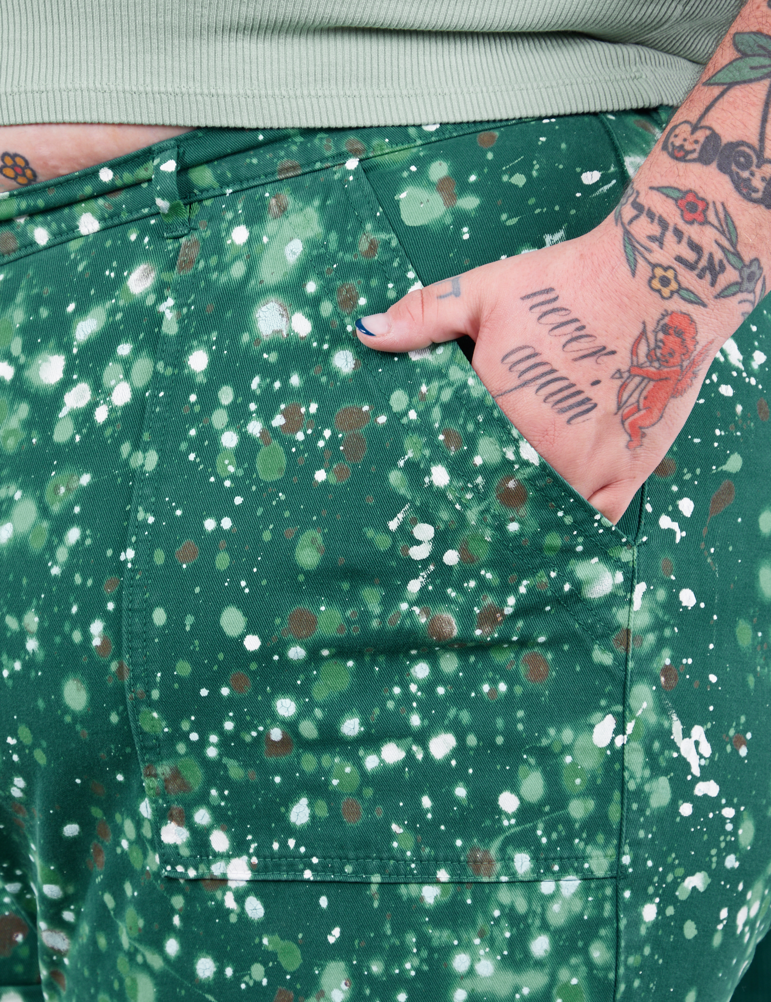 Front pocket close up of Marble Splatter Work Pants in Hunter Green. Sam has their hand in the pocket.